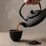 Main tenant une théière en fonte Tetsubin noire pour verser du thé chaud fumant dans une tasse assortie sur une table en pierre.